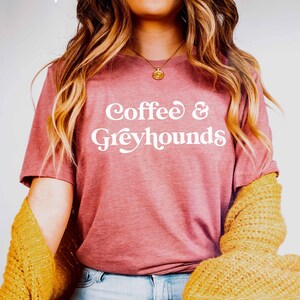 May include: A woman wearing a maroon t-shirt with the text "Coffee & Greyhounds" in white. She is wearing blue jeans and a yellow cardigan.