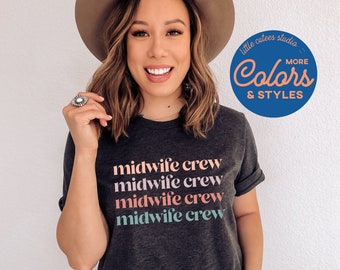 May include: A dark grey t-shirt with the text "midwife crew" repeated four times in different pastel colours. The text is stacked vertically on the shirt. The shirt is being worn by a person with brown hair and a brown hat. The background is a white wall. The image also includes a blue circle with the text "little cutes studio Colors & Styles" in white.