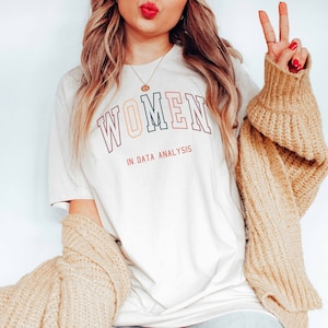 May include: A white t-shirt with the text "WOMEN IN DATA ANALYSIS" in rainbow colored letters. The person wearing the shirt is also wearing a brown cardigan and blue jeans.