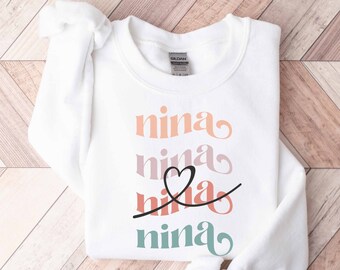 May include: White sweatshirt with the name "nina" written four times in different colors and styles. The name is stacked vertically with a heart symbol between the second and third "nina".
