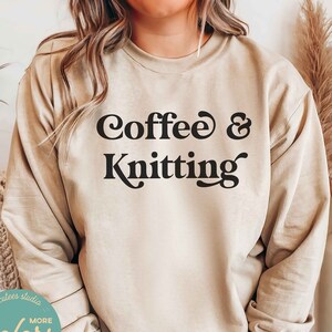May include: A beige sweatshirt with the text "Coffee & Knitting" in black retro-style font. The sweatshirt is worn by a person and the text is centered on the chest.