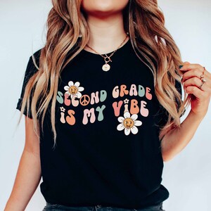 Puede incluir: Camiseta negra con la frase "Second Grade is My Vibe" en letras coloridas de estilo retro. El diseño incluye margaritas con caras sonrientes y un símbolo de la paz, creando un tema alegre de regreso a la escuela.