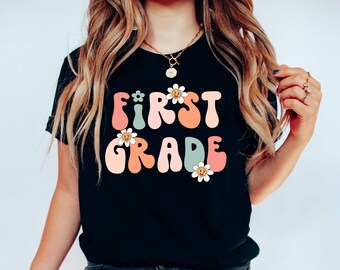 May include: A black t-shirt with the text "FIRST GRADE" in colorful, retro-style letters with daisies.