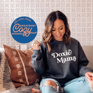 May include: A woman wearing a dark grey sweatshirt with the text "Doxie Mama" in white. She is sitting on a sofa with a brown and white patterned throw pillow.
