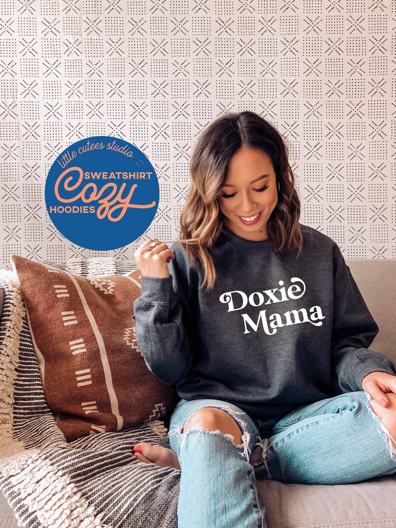May include: A woman wearing a dark grey sweatshirt with the text "Doxie Mama" in white. She is sitting on a sofa with a brown and white patterned throw pillow.