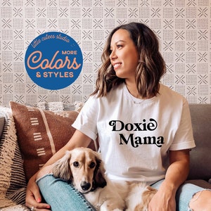 May include: A woman wearing a white t-shirt with the text "Doxie Mama" in black lettering, sitting on a sofa with a small, light brown dachshund dog in her lap. The woman is smiling and looking to the side. The background is a white wall with a black and white geometric pattern.