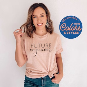 May include: A woman wearing a light pink t-shirt with the text "FUTURE engineer" in black script. The shirt is tied at the waist. She is wearing blue jeans and has long brown hair. The image also includes a blue circle with the text "little cutees studio... Colors MORE & STYLES" in white.