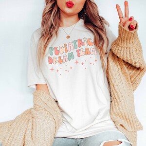 May include: A white t-shirt with the text "Pediatric Dream Team" in colorful, retro-style lettering. The shirt features small, sparkly stars.