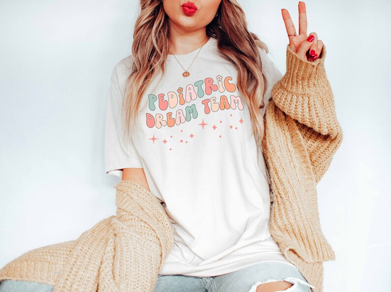 May include: A white t-shirt with the text "Pediatric Dream Team" in colorful, retro-style lettering. The shirt features small, sparkly stars.