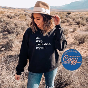 May include: A black sweatshirt with white text that reads "eat. sleep. meditation. repeat." The sweatshirt is being worn by a person in a desert setting. The text "little cutes studio" and "Cozy" are visible on a blue circle.