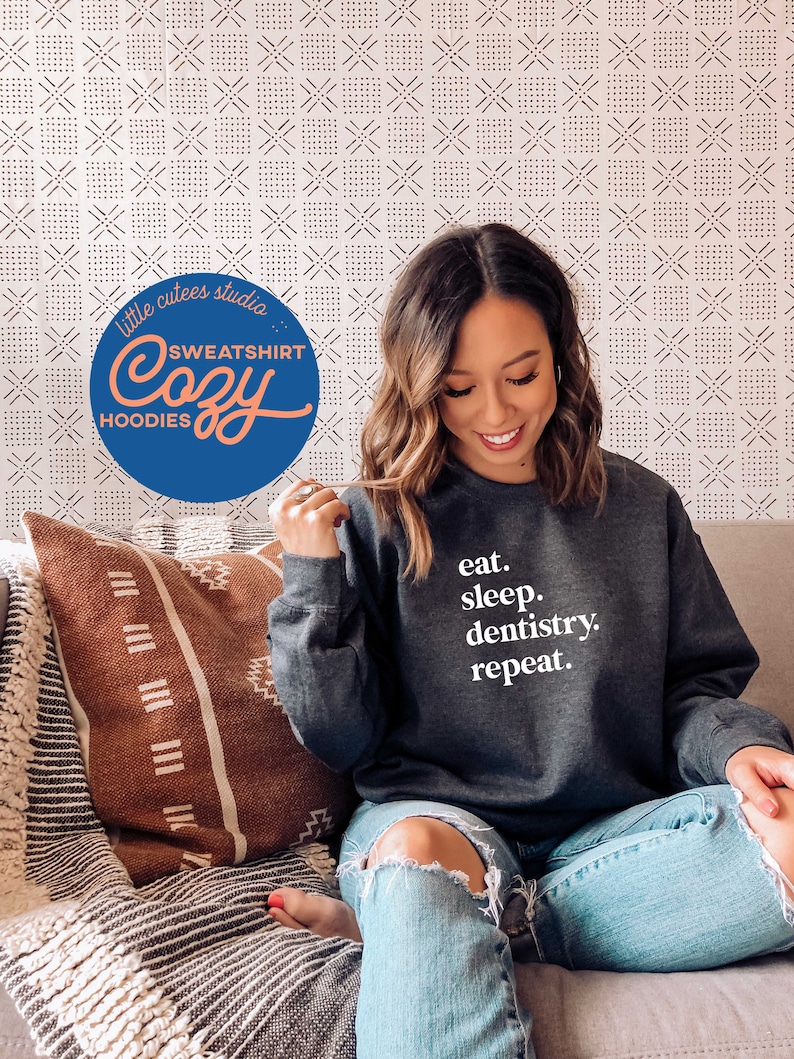 May include: Dark gray sweatshirt with the text "eat. sleep. dentistry. repeat." in white. The sweatshirt is part of a cozy hoodies collection. The image also includes a decorative pillow and a patterned background.