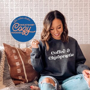 May include: A woman wearing a dark gray sweatshirt with the text "Coffee & Chiropractic" in white. She is sitting on a couch with a brown and white patterned throw pillow.