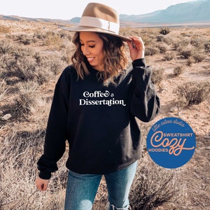 May include: A black sweatshirt with the text "Coffee & Dissertation" in white lettering. The sweatshirt is being worn by a person in a desert setting. The image also includes a logo for "little cutes studio" with the text "Cozy Sweatshirt Hoodies" in a peach color.