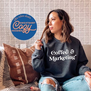 May include: A woman wearing a dark gray sweatshirt with the text "Coffee & Marketing" in white. She is sitting on a couch with a brown and white patterned throw pillow.