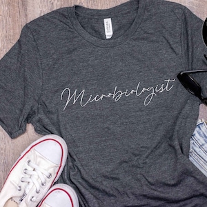 May include: Dark gray t-shirt with the word "Microbiologist" in a cursive font. The shirt is paired with white sneakers, sunglasses, and denim shorts. A black circle with white text reads "more colors & more styles."