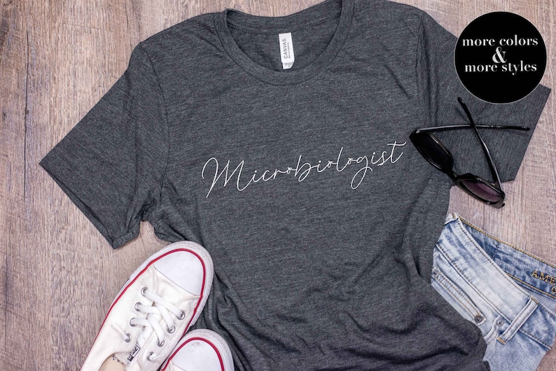 May include: Dark gray t-shirt with the word "Microbiologist" in a cursive font. The shirt is paired with white sneakers, sunglasses, and denim shorts. A black circle with white text reads "more colors & more styles."