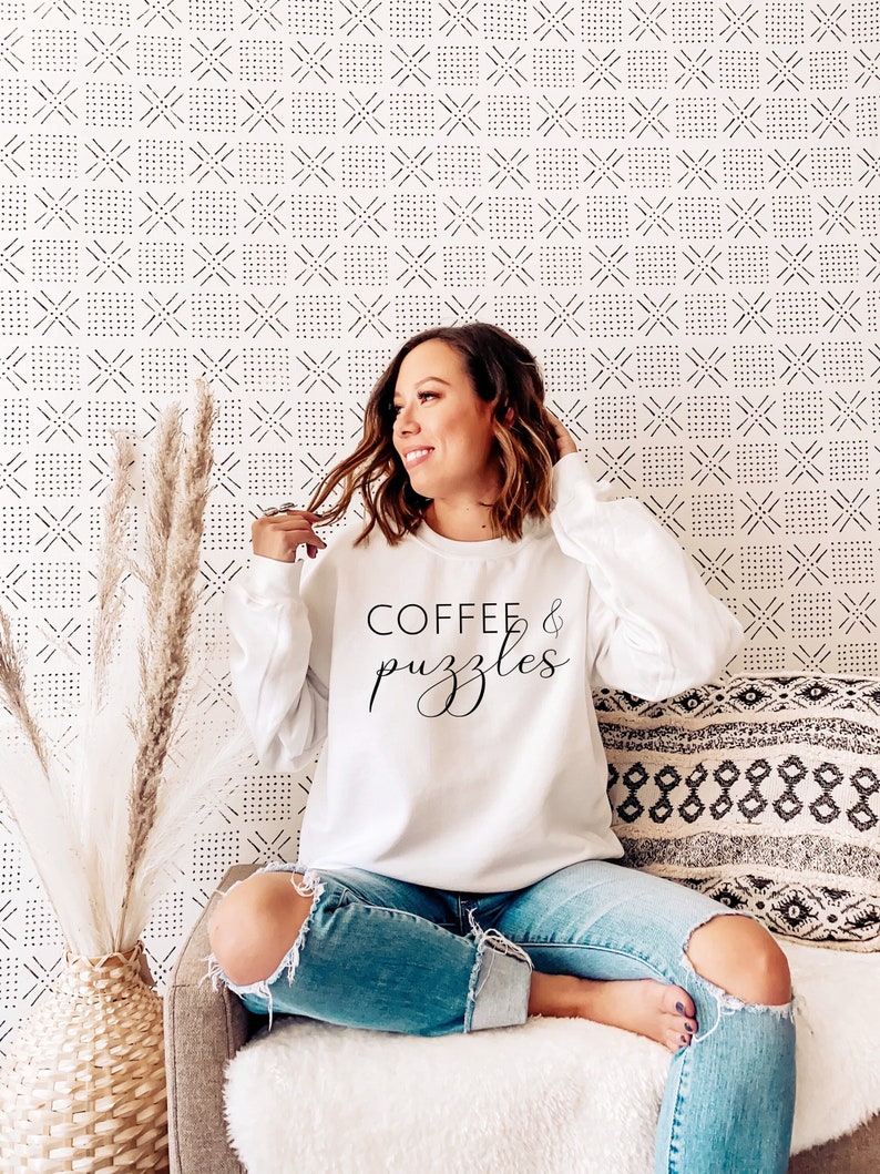 May include: A white sweatshirt with the text "COFFEE & puzzles" in black lettering. The sweatshirt is being worn by a person sitting on a couch.