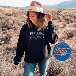 May include: A black sweatshirt with the text "FUTURE professor" in white script. The sweatshirt is being worn by a person in a desert setting. The image also includes a logo for "little cutes studio" with the text "SWEATSHIRT Cozy HOODIES".