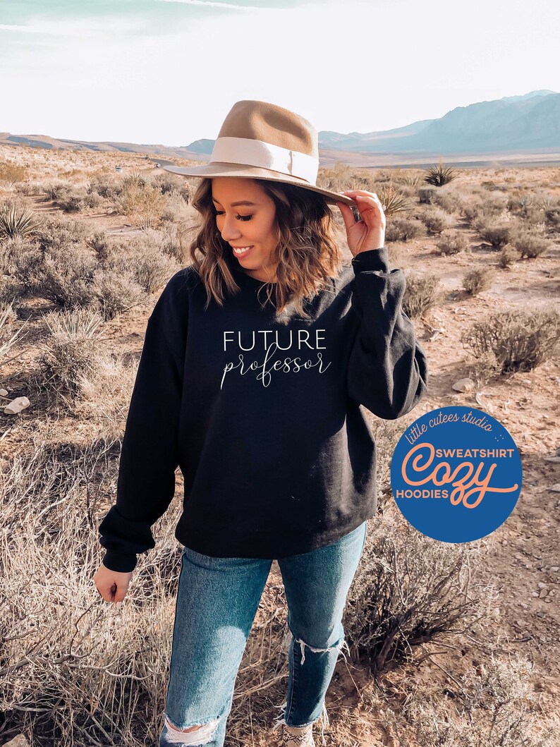 May include: A black sweatshirt with the text "FUTURE professor" in white script. The sweatshirt is being worn by a person in a desert setting. The image also includes a logo for "little cutes studio" with the text "SWEATSHIRT Cozy HOODIES".