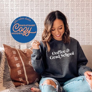 May include: A dark gray sweatshirt with the text "Coffee & Grad School" printed in white. The sweatshirt is being worn by a person sitting on a couch.
