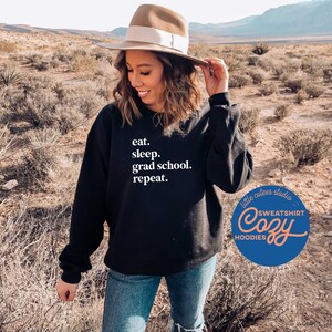 May include: A black sweatshirt with white text that reads "eat. sleep. grad school. repeat." The sweatshirt is being worn by a person in a desert setting. The text "little cutees studio" and "Cozy" are visible on a blue circle with white text.