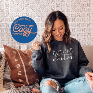 May include: A dark gray sweatshirt with the words "FUTURE doctor" in white script. The sweatshirt is part of a cozy collection, as indicated by the text in a circular logo. The image shows a person wearing the sweatshirt.