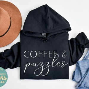 May include: Black hooded sweatshirt with the text "COFFEE & puzzles" in white lettering. The sweatshirt is laid flat on a white surface with a brown hat and blue jeans.