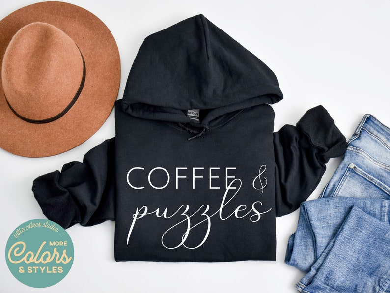 May include: Black hooded sweatshirt with the text "COFFEE & puzzles" in white lettering. The sweatshirt is laid flat on a white surface with a brown hat and blue jeans.