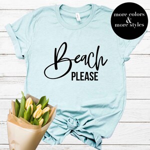 May include: A light blue t-shirt with the text "Beach Please" in black cursive lettering. The shirt is knotted at the bottom and has rolled up sleeves.