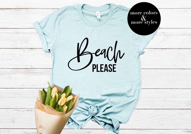 May include: A light blue t-shirt with the text "Beach Please" in black cursive lettering. The shirt is knotted at the bottom and has rolled up sleeves.