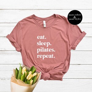 May include: A dusty rose t-shirt with the text "eat. sleep. pilates. repeat." printed in white.