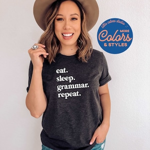 May include: A dark gray t-shirt with white text that reads "eat. sleep. grammar. repeat." The model is wearing the shirt and a brown hat. The background is a white wall with a blue and pink logo that reads "little cutes studio Colors & Styles".