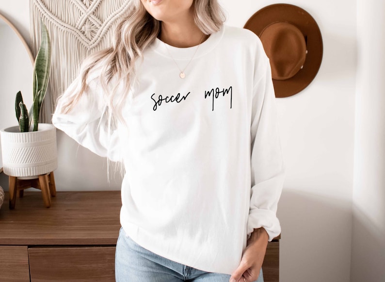 May include: White long-sleeved sweatshirt with the text "Soccer Mom" written in black cursive script.