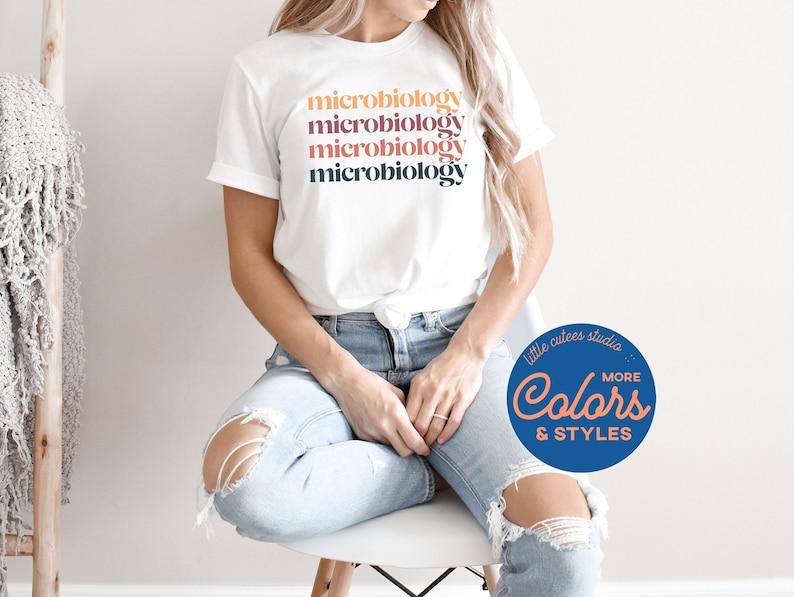 May include: A white t-shirt with the word "microbiology" repeated five times in different colors. The colors are orange, pink, purple, and gray. The shirt is being worn by a person sitting on a chair. The image also includes a blue circle with the text "little cutes studio" and "MORE Colors & STYLES" in white.