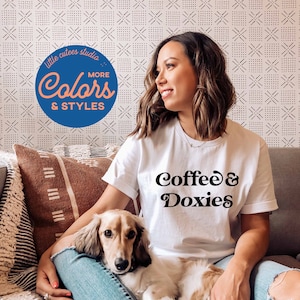 Puede incluir: Una mujer con una camiseta blanca con el texto "Coffee & Doxies" en letras negras, sentada en un sof&aacute; con un peque&ntilde;o perro dachshund marr&oacute;n claro y blanco en su regazo. El sof&aacute; tiene un coj&iacute;n de tiro con estampado marr&oacute;n y blanco.