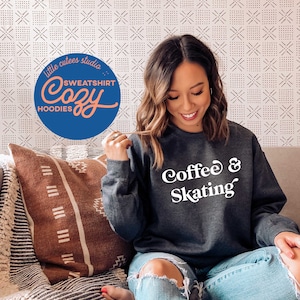 May include: A woman wearing a dark gray sweatshirt with the text "Coffee & Skating" in white. She is sitting on a couch with a brown and white patterned throw pillow.