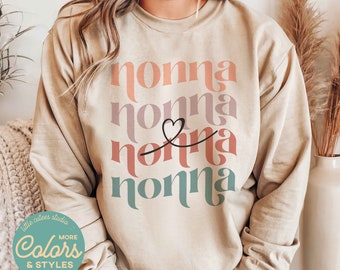 May include: A beige sweatshirt with the word "nonna" repeated four times in different colors and fonts. The word "nonna" is stacked vertically with a small heart between the second and third words. The sweatshirt has a crew neck and long sleeves.
