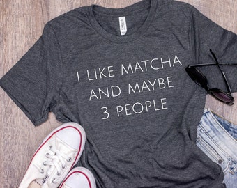 May include: A dark gray t-shirt with the text "I like matcha and maybe 3 people" printed in white. The shirt is laid flat on a wood surface with a pair of sunglasses and a pair of white sneakers.