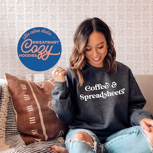 May include: A dark gray sweatshirt with the text "Coffee & Spreadsheets" in white. The sweatshirt is being worn by a person sitting on a couch with a brown and white patterned throw pillow.