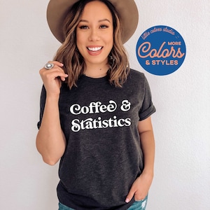 May include: A dark gray t-shirt with the text "Coffee & Statistics" in white. The shirt is being worn by a person with a brown hat. A blue circle with the text "little cutes studio Colors & STYLES" is in the upper right corner of the image.