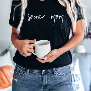 May include: A woman wearing a black t-shirt with the text "soccer mom" in white script. She is holding a white ceramic mug.