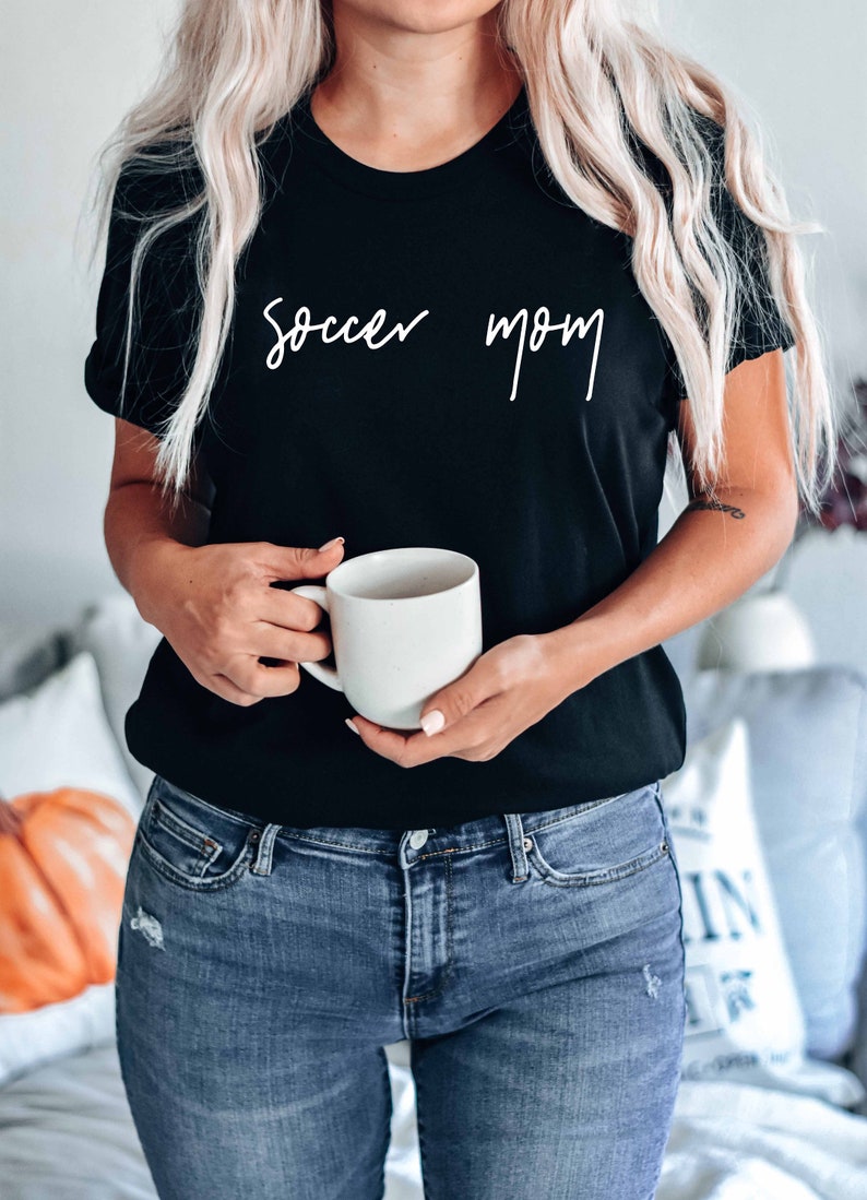May include: A woman wearing a black t-shirt with the text "soccer mom" in white script. She is holding a white ceramic mug.