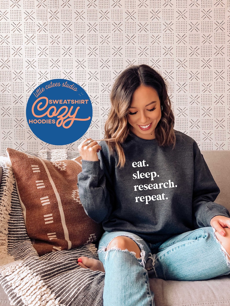 May include: A woman wearing a dark gray sweatshirt with the text "eat. sleep. research. repeat." on it. She is sitting on a couch with a brown and white patterned throw pillow.