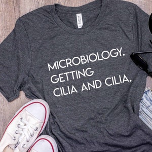 May include: A dark gray t-shirt with the text "MICROBIOLOGY. GETTING CILIA AND CILIA." printed in white.