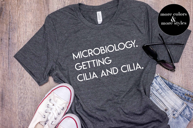 Pu&ograve; includere: Una maglietta grigio scuro con il testo "MICROBIOLOGY. GETTING CILIA AND CILIA." stampato in bianco.