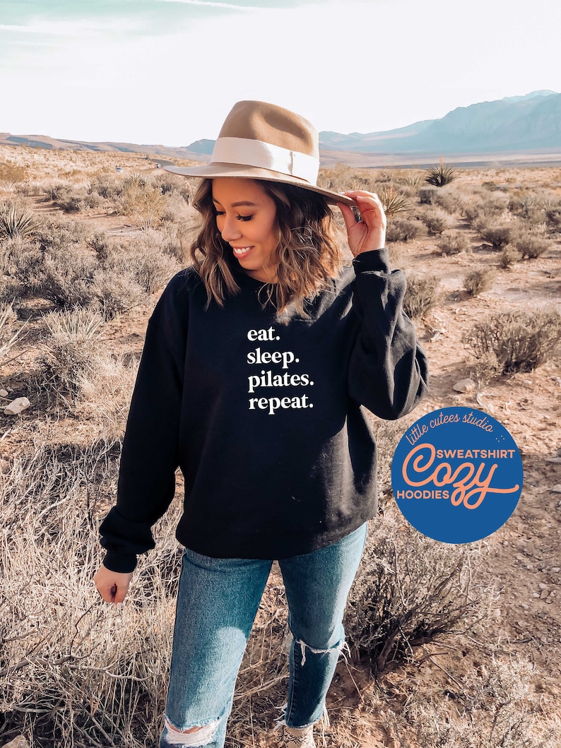 May include: A black sweatshirt with the text "eat. sleep. pilates. repeat." in white. The sweatshirt is being worn by a person in a desert setting. The image also includes a logo for "little cutes studio" with the text "SWEATSHIRT Cozy HOODIES".