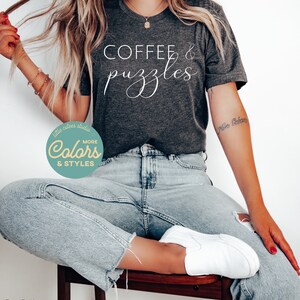 May include: A dark heather grey t-shirt with the text "COFFEE & puzzles" in white cursive lettering. The shirt is being worn by a person sitting on a wooden stool. The person is wearing light blue jeans and white sneakers.
