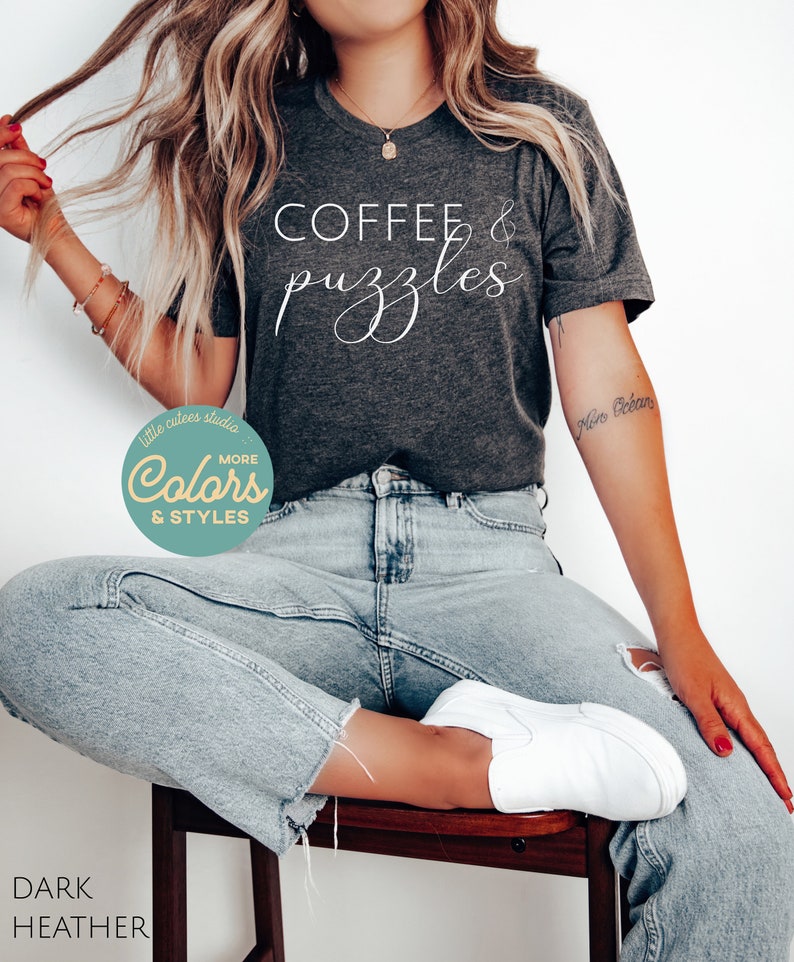May include: A dark heather grey t-shirt with the text "COFFEE & puzzles" in white cursive lettering. The shirt is being worn by a person sitting on a wooden stool. The person is wearing light blue jeans and white sneakers.