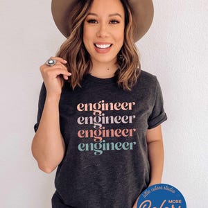 May include: Dark gray t-shirt with the word "engineer" printed in pink, peach, and teal. The shirt has rolled-up sleeves and is paired with a brown hat. The shirt also has the text "little cutees studio" and "Colors & Styles" on it.