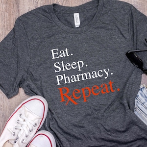 May include: A dark gray t-shirt with the text "Eat. Sleep. Pharmacy. Repeat." in white and red lettering. The shirt is laid flat on a wooden surface with a pair of sunglasses, a pair of white sneakers, and a pair of blue jeans.